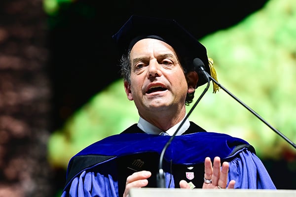 Dean Brian T. Edwards in blue academic gown and cap speaks at a podium with microphones.