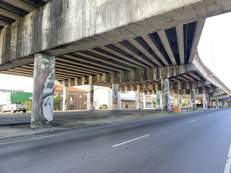 Concrete highway overpass with tree murals on pillars, above a road and buildings.