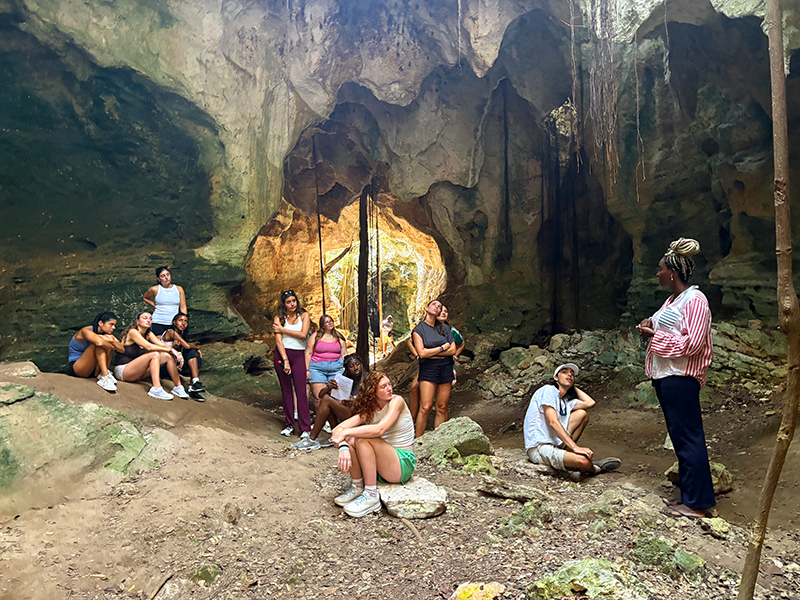 Photograph: Diverse group gathers in a large, earthy cave with golden light from an opening.
