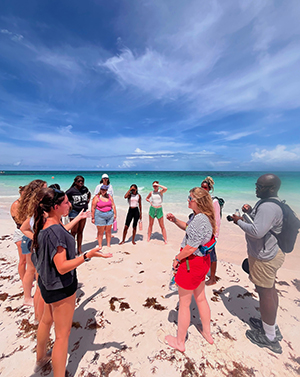 Group of people on a sunny beach with turquoise water, some listening, one holding a camera.
