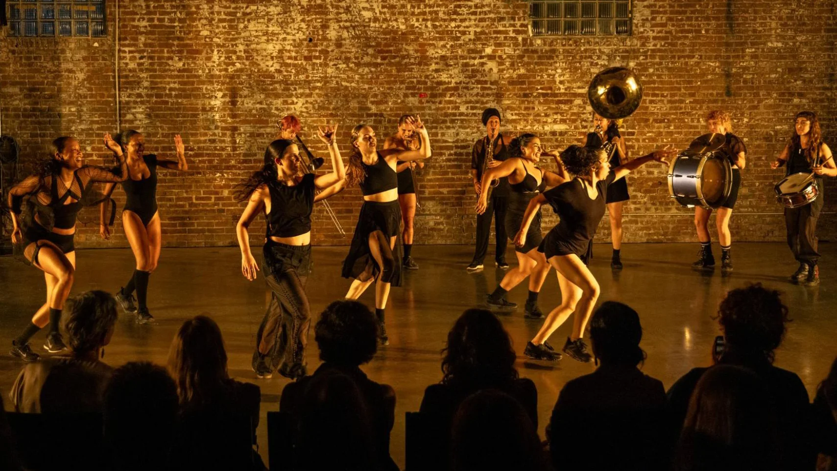 Dancers and musicians perform on stage for an audience, warm brick wall background.
