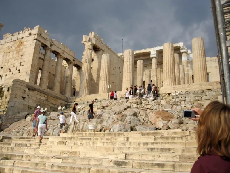 Tourists climb steps to the ancient Propylaea on the Acropolis under a cloudy sky.