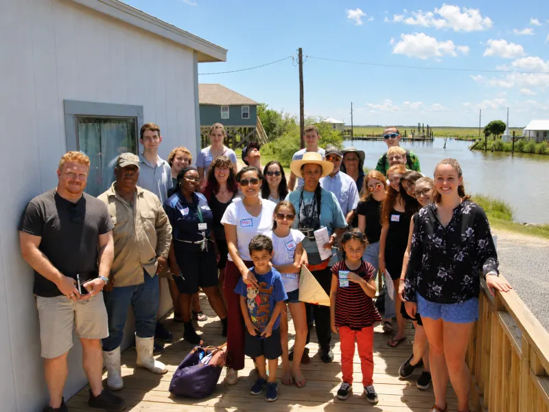 Diverse group of adults and children on a wooden deck beside a white building and a waterway.