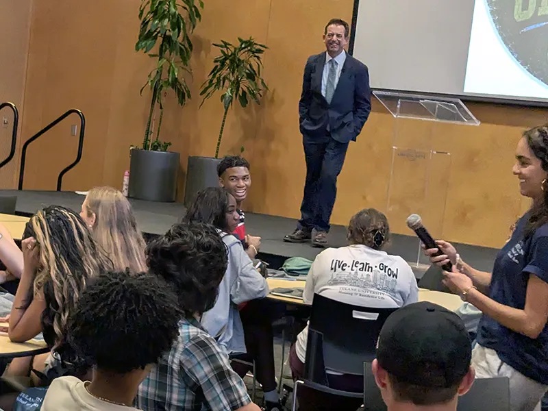 Speaker on stage addresses diverse group of students; woman with mic facilitates.