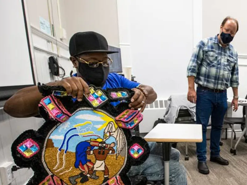 Man in mask holds a vibrant, round beaded artwork with a central scene of figures; another man obser