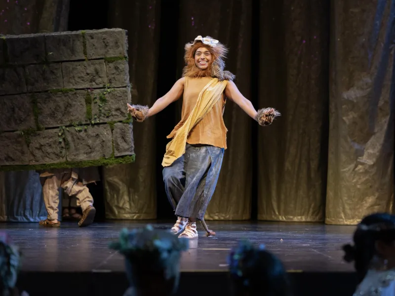 Smiling person in lion costume on stage, dancing near a large gray block.